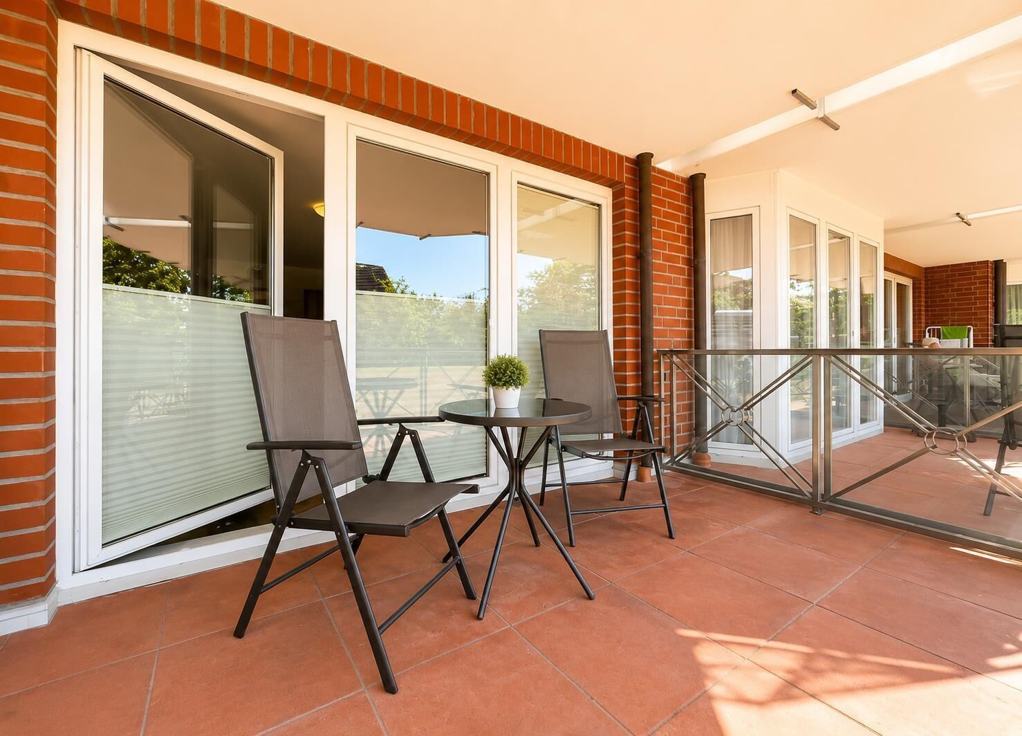 Eine Terrasse mit zwei Stühlen und einem Tisch, einem Glasgeländer und Blick auf einen Garten durch große Fenster.