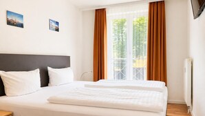Ein Hotelzimmer mit einem Bett, zwei Kissen, einem Kopfteil, einem Fenster mit Vorhängen, einem Heizkörper und zwei Wandbildern.