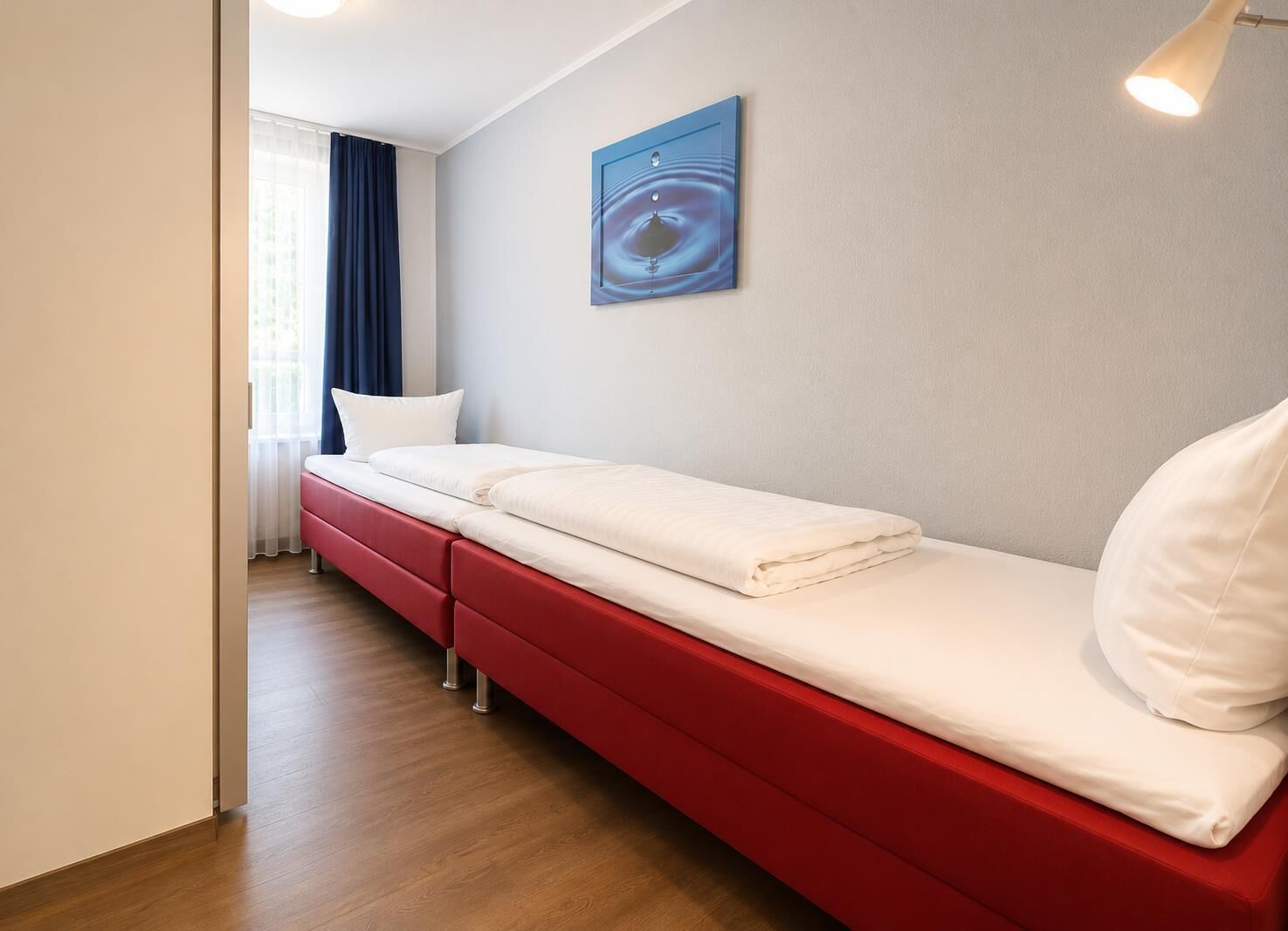 Habitación de hotel con una cama roja, ropa de cama blanca, una obra de arte azul en la pared y una ventana con cortinas.