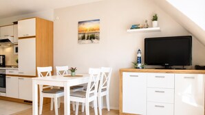 A modern kitchen with white cabinets, a dining table with chairs, a television, and a small shelf with books and a plant. - Haus am Mühlenpark Whg. 14, Steinmarnerstr.77/79, 27476 Cuxhaven (Cuxhaven- Döse)