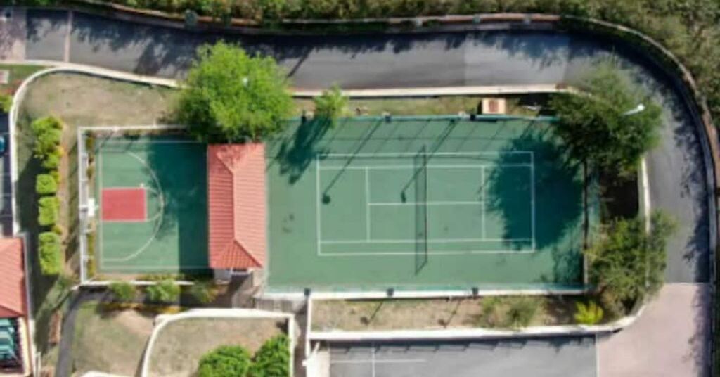 An aerial view of a tennis court with a red roof, surrounded by greenery and a curved road.