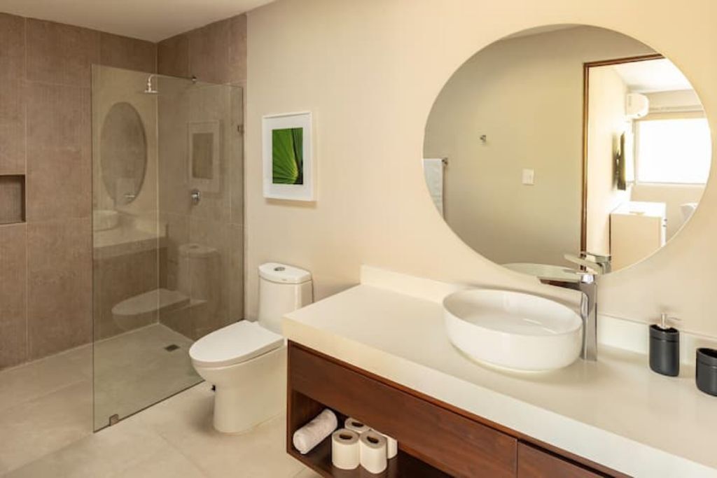 A modern bathroom with a glass-enclosed shower, a white toilet, a large round mirror, and a white sink.