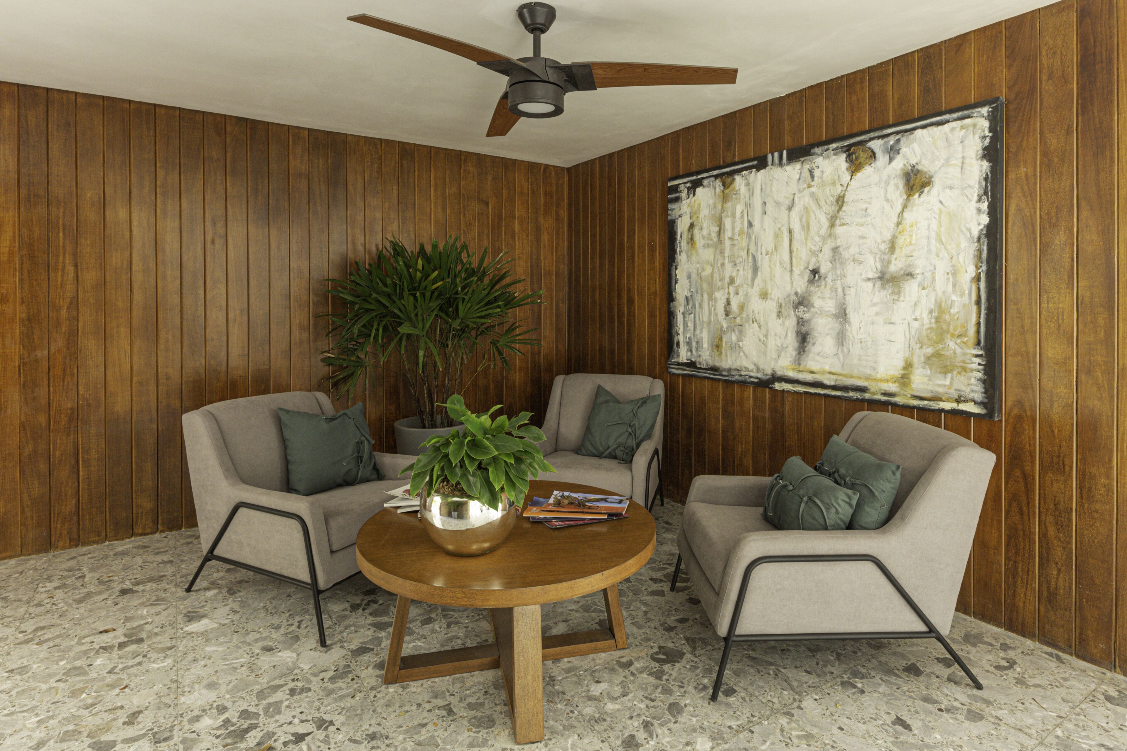 Una habitación con paredes de madera, ventilador de techo, dos sillones, una mesa redonda con una planta y un cuadro abstracto grande.