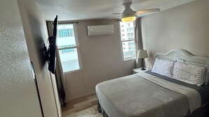 Quarto com cama, ventoinha no teto, ar condicionado, janela e mesa de cabeceira com abajur.