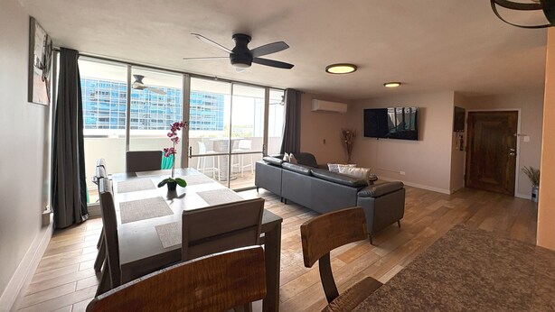 Uma sala de estar ampla, com uma mesa de jantar, cadeiras, um sofá e uma TV fixada na parede.