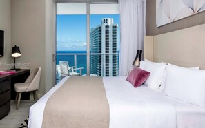 A hotel room with a large bed, a desk, a chair, and a view of a tall building and the ocean. - Luxury Apartment  in Hollywood Beach (Hollywood)