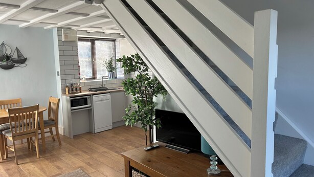 A modern kitchen with white cabinets, a built-in oven, and a sink. There is a dining area with wooden chairs and a table. A large plant is in the corner, and a TV is on a wooden stand. - Barn conversion overlooking the golf course to the sea (Bude)