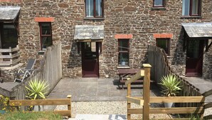 A stone building with multiple windows and doors, a wooden fence, and a paved patio area with potted plants. - Barn conversion overlooking the golf course to the sea (Bude)