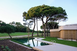 A modern outdoor pool area with a wooden fence, a concrete pathway, and a large tree. - Marinha Prime (Cascais)