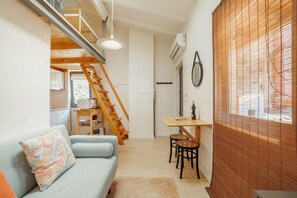 Un espacio habitable pequeño con sofá, una mesa de comedor con sillas, una escalera de madera y una ventana con persianas.