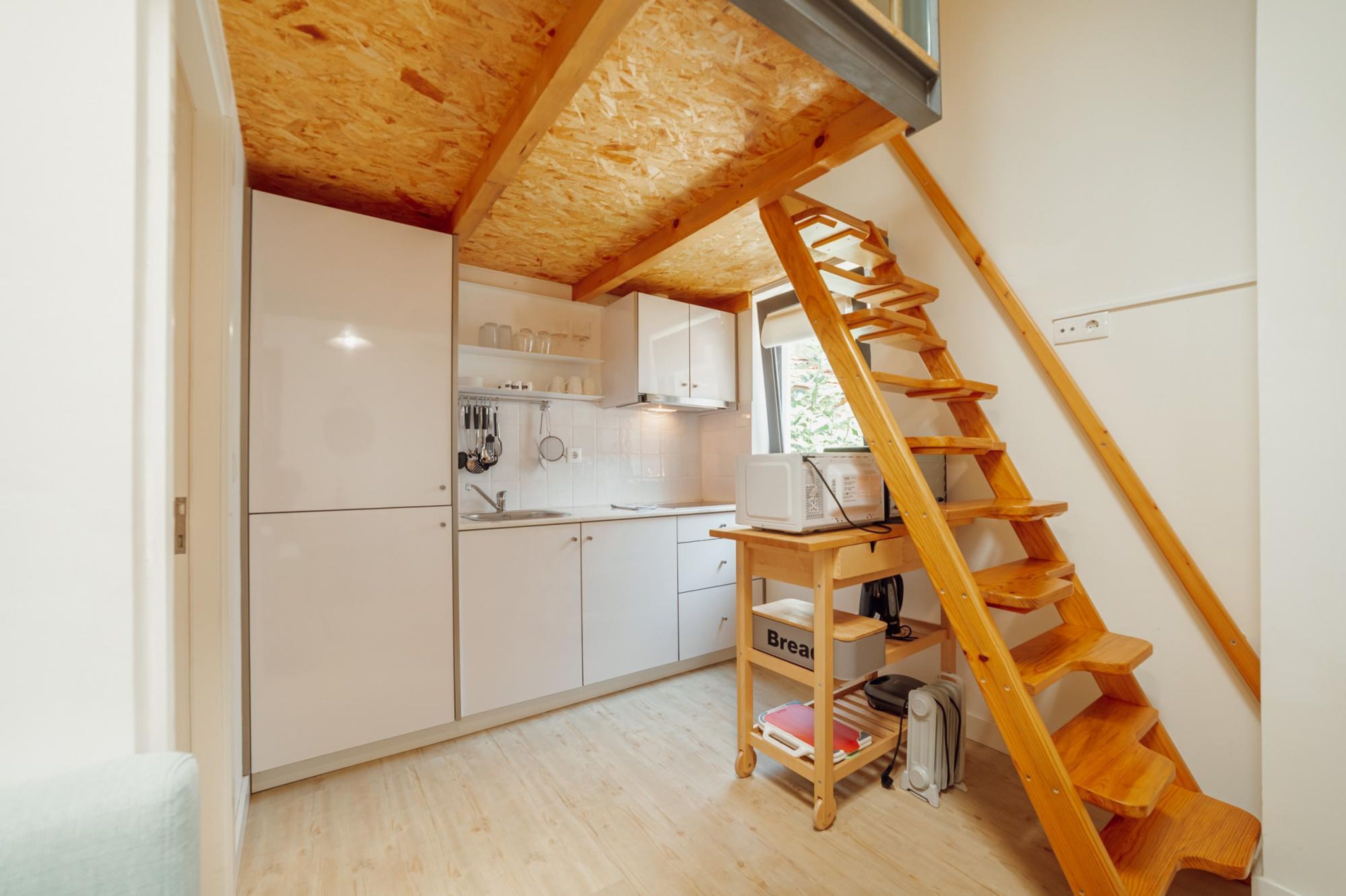 Cocina compacta con armarios blancos, una escalera de madera y una pequeña mesa que dice 'Pan'.