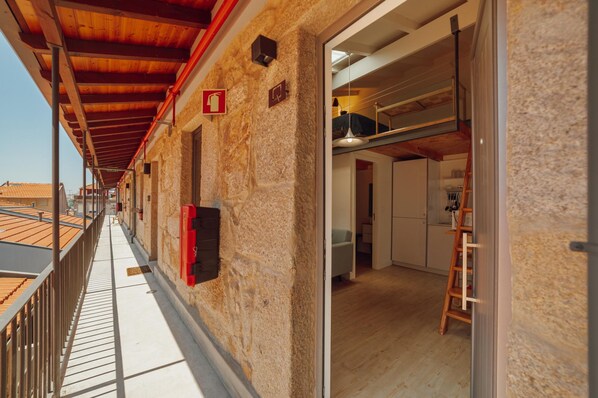 Un pasillo con pared de piedra, techo de madera y vista a una habitación con cama elevada y escalera.