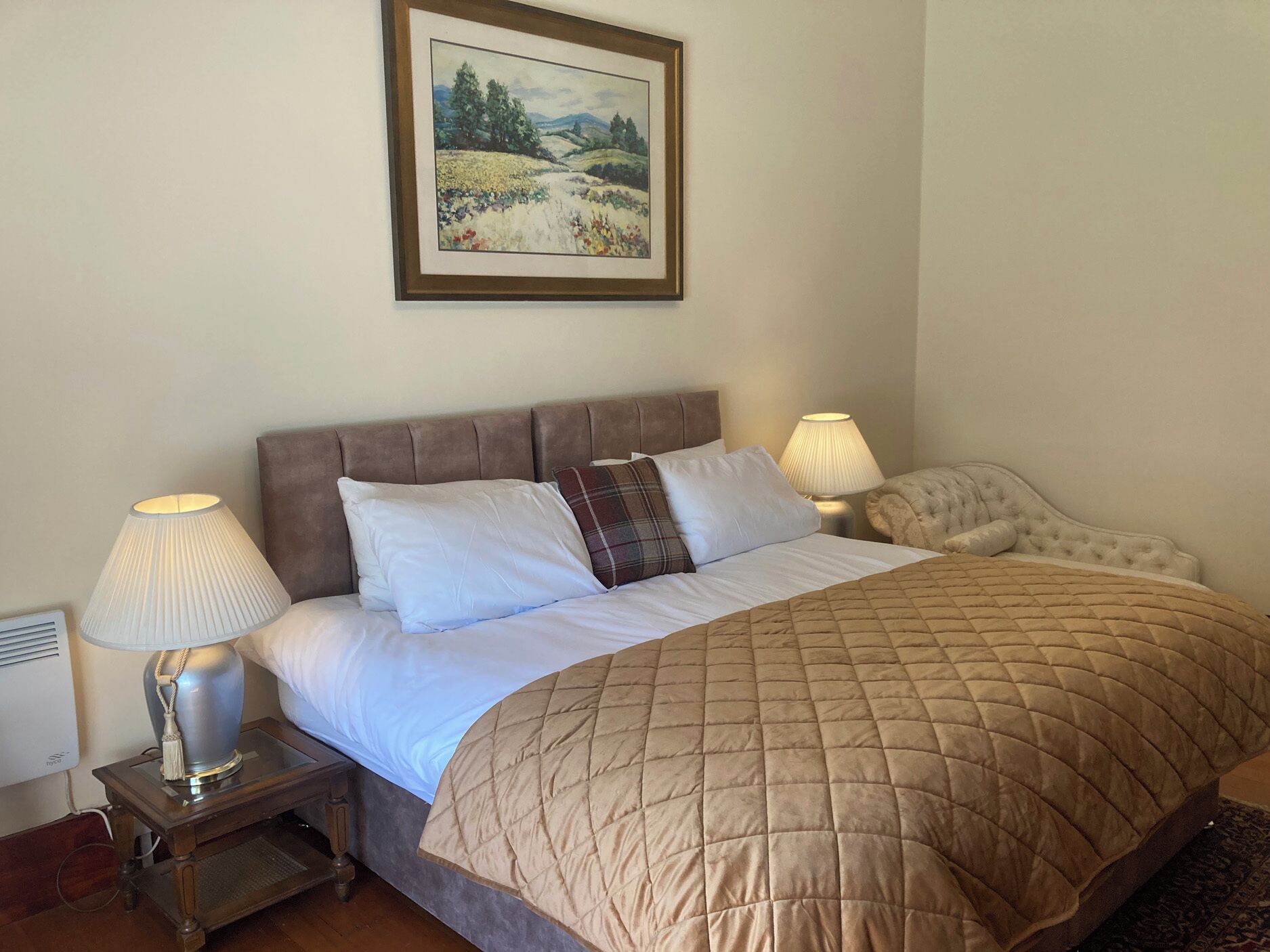A bedroom with a bed, two bedside lamps, a framed landscape painting, and a radiator.