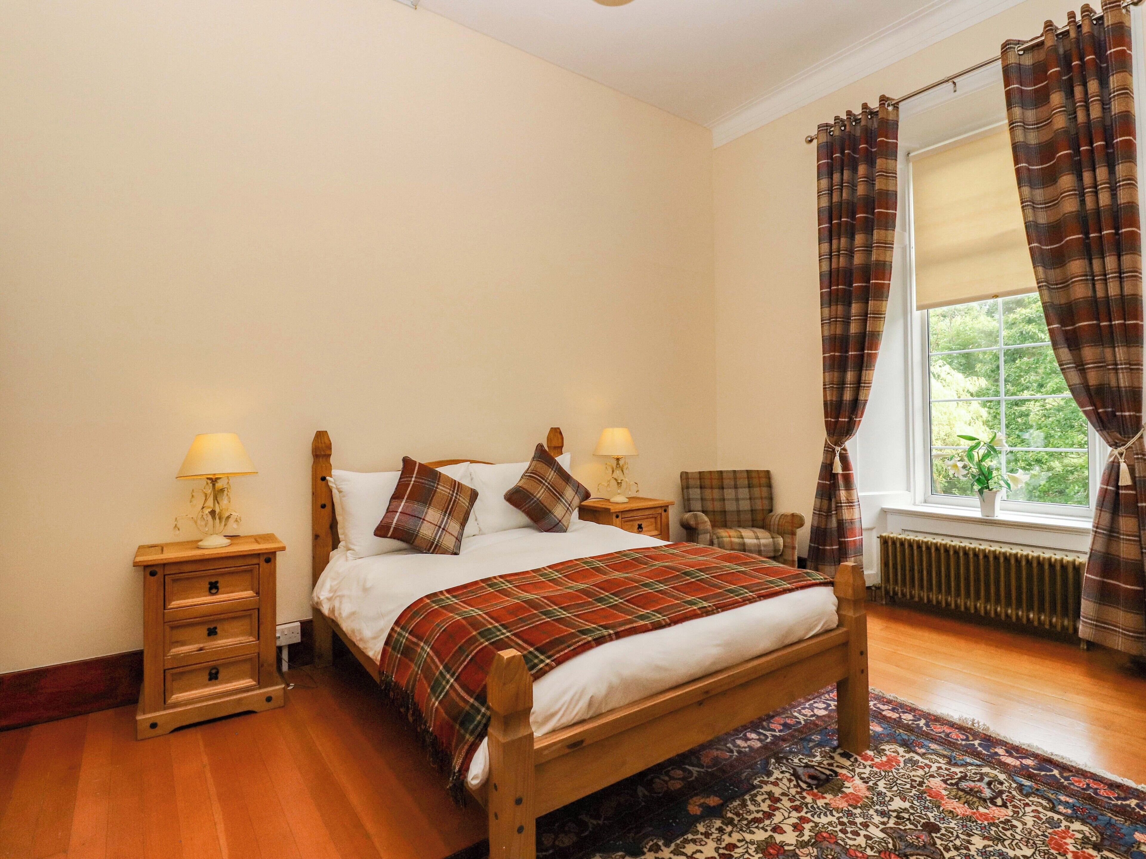 A bedroom with a wooden bed, two bedside tables with lamps, a chair, a window with curtains, and a radiator.