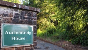 A sign for 'Auchenroig House' mounted on a brick wall beside a paved path surrounded by greenery. - Rob Roy Apartment (Glasgow)