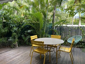 A round wooden table with yellow metal chairs and a blue woven chair, surrounded by lush tropical plants and a bamboo fence.