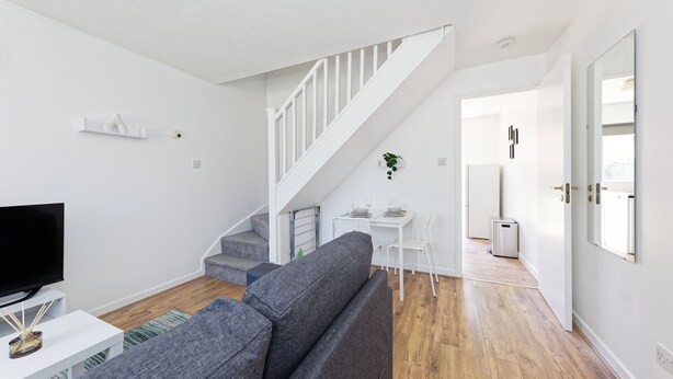 A modern living room with a grey sofa, a flat-screen TV, and a staircase leading to another room. - Lovely 2-bed House in Kent - Parking Available (Greenhithe)
