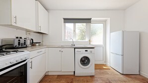 A compact kitchen with white cabinets, a built-in washing machine, a stove, and a refrigerator. - Lovely 2-bed House in Kent - Parking Available (Greenhithe)