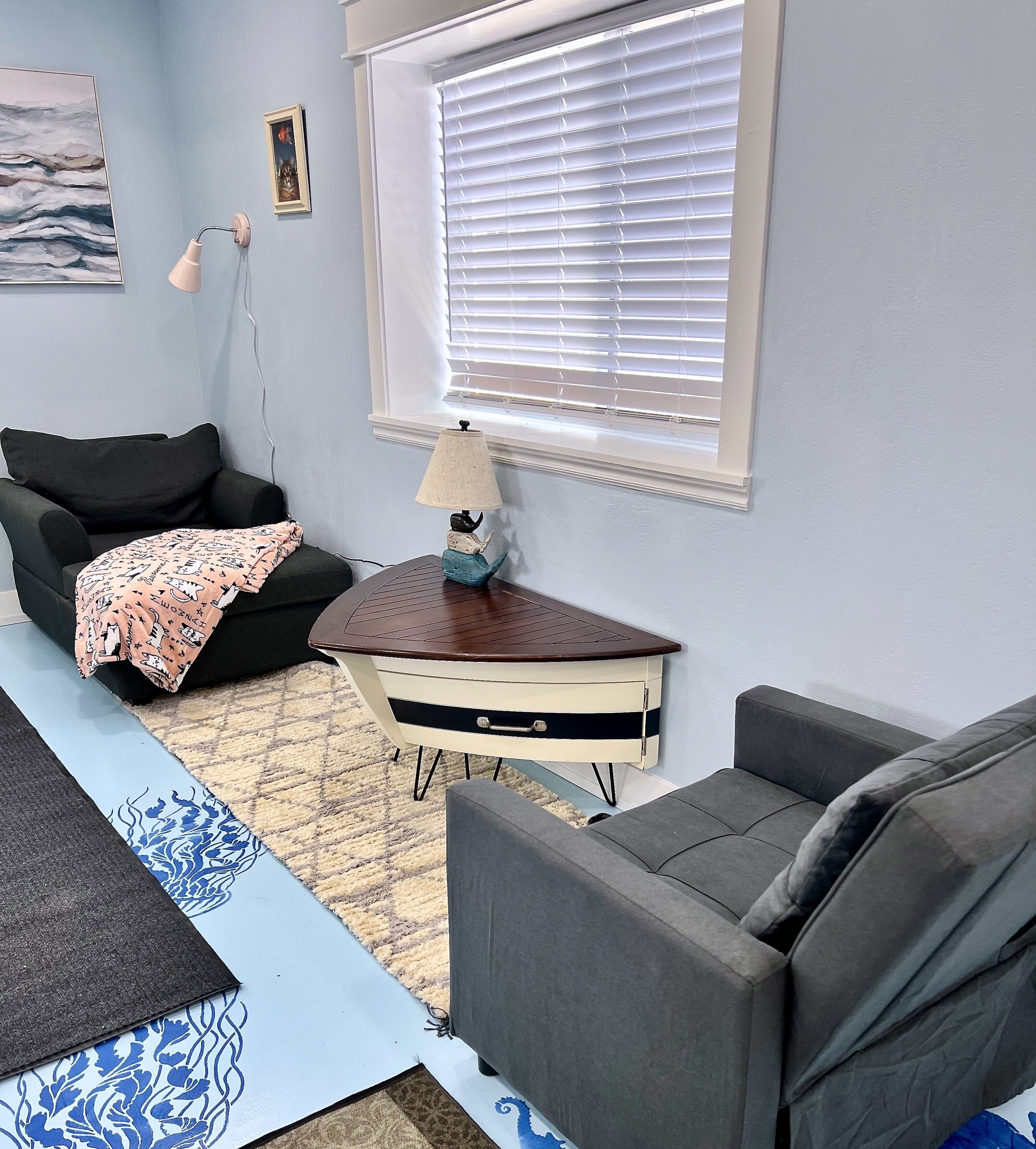 A living room with a grey sofa, a small table with a lamp, a window with blinds, and a patterned rug.