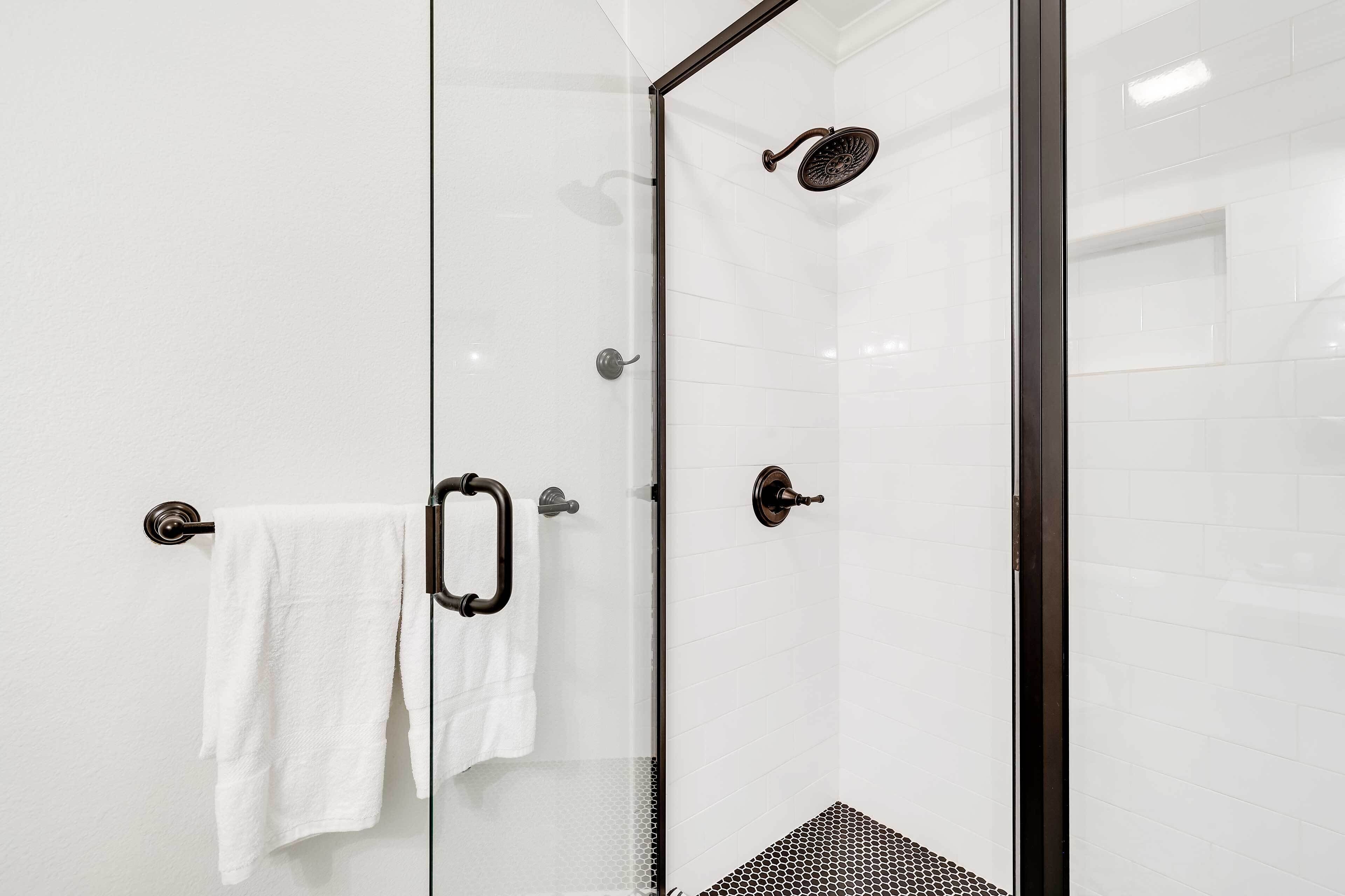 A glass-enclosed shower with a white tiled wall, a towel hanging on a metal towel rack, and a black textured shower floor.