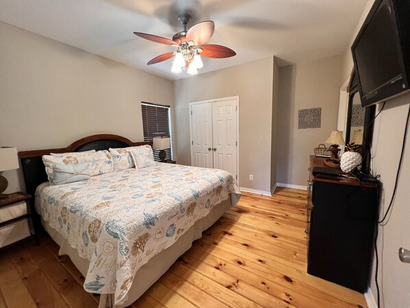 A bedroom with a bed, a ceiling fan, a television, and a wooden floor.