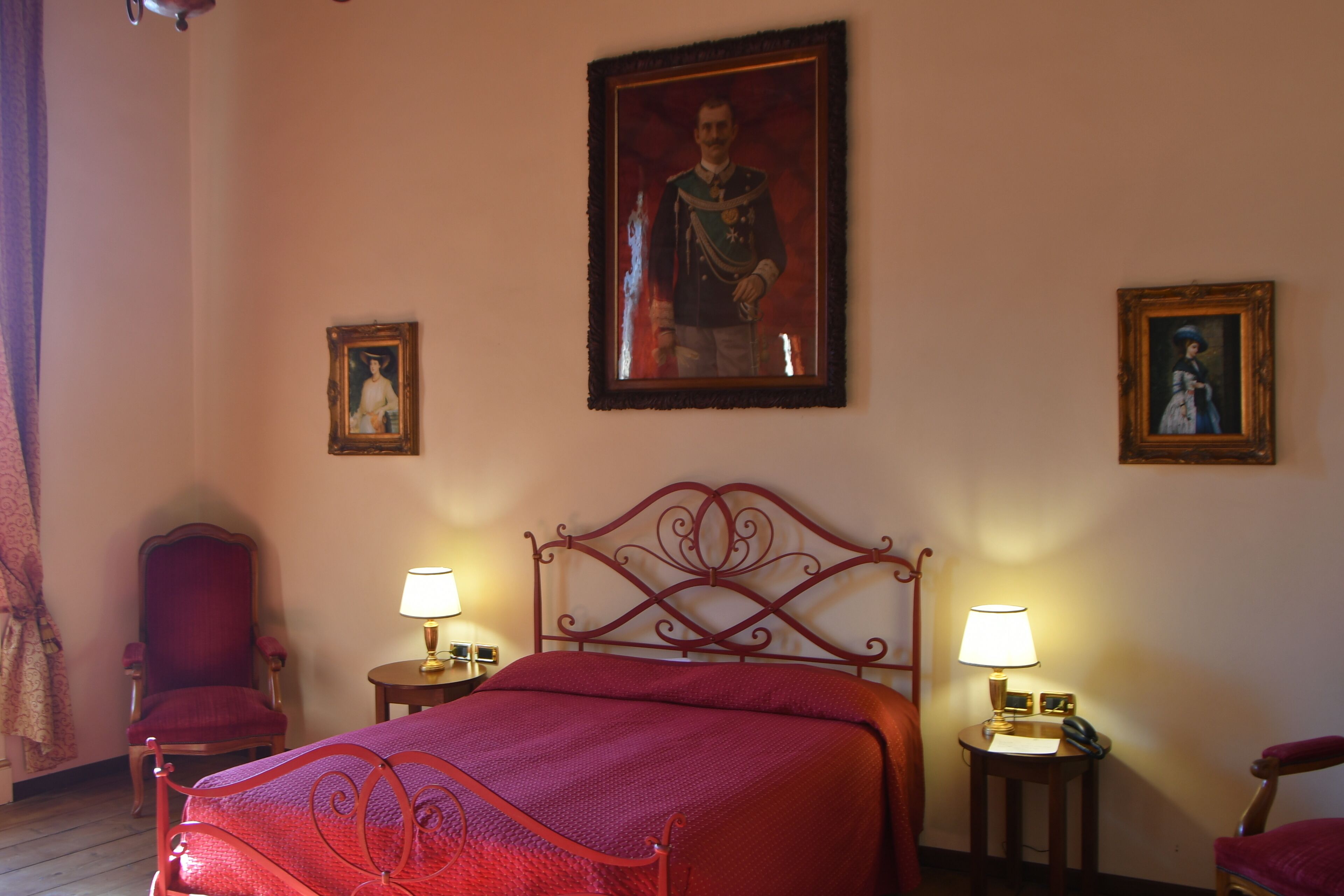Une chambre d’hôtel avec un lit en métal dont le cadre est rouge, deux tables de chevet avec des lampes, un grand portrait encadré accroché au mur et deux chaises.