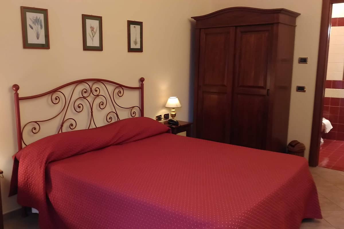 A neatly made bed with a red bedspread, a wooden headboard, and a nightstand with a lamp. There are framed pictures on the wall above the bed.