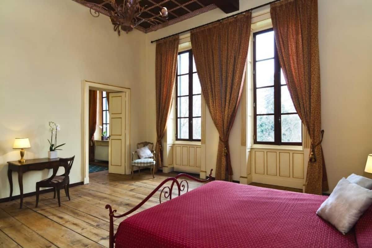 A bedroom with a wooden floor, a bed with a red bedspread, a desk with a lamp, and a chair.