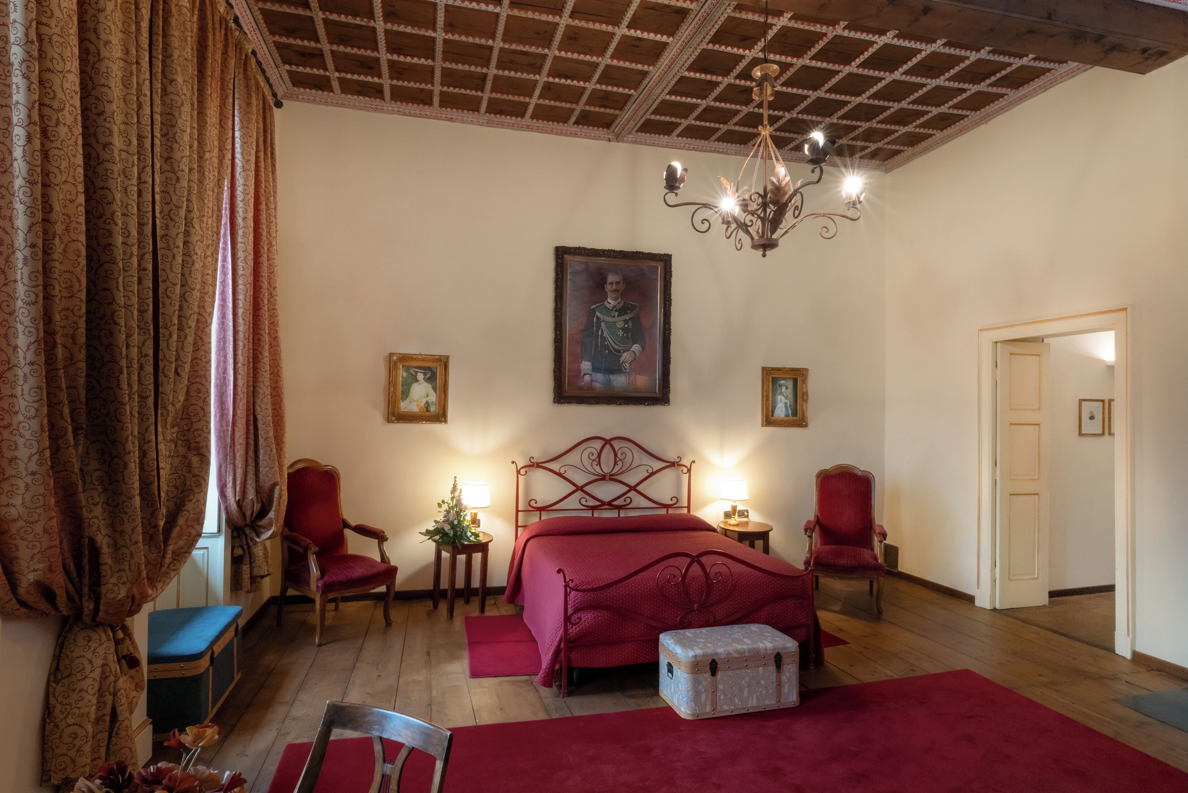 Un dormitorio con techo de madera, una cama roja, dos sillas rojas, un banco, una mesita de noche con una lámpara y un cuadro en la pared.