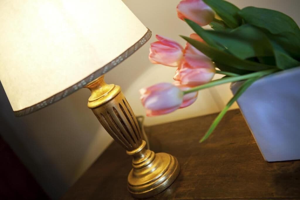 Une lampe de table avec un abat-jour blanc et un pied doré, posée sur une surface en bois à côté d’un vase contenant des tulipes roses.