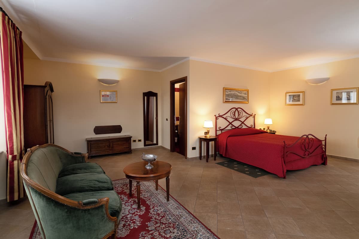 A hotel room with a bed, a green armchair, a small table with a lamp, a dresser, and a mirror.