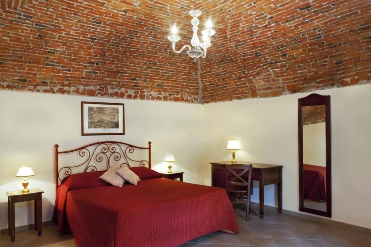 A room with a brick ceiling, a bed with a red bedspread, two bedside tables with lamps, a desk, and a mirror.