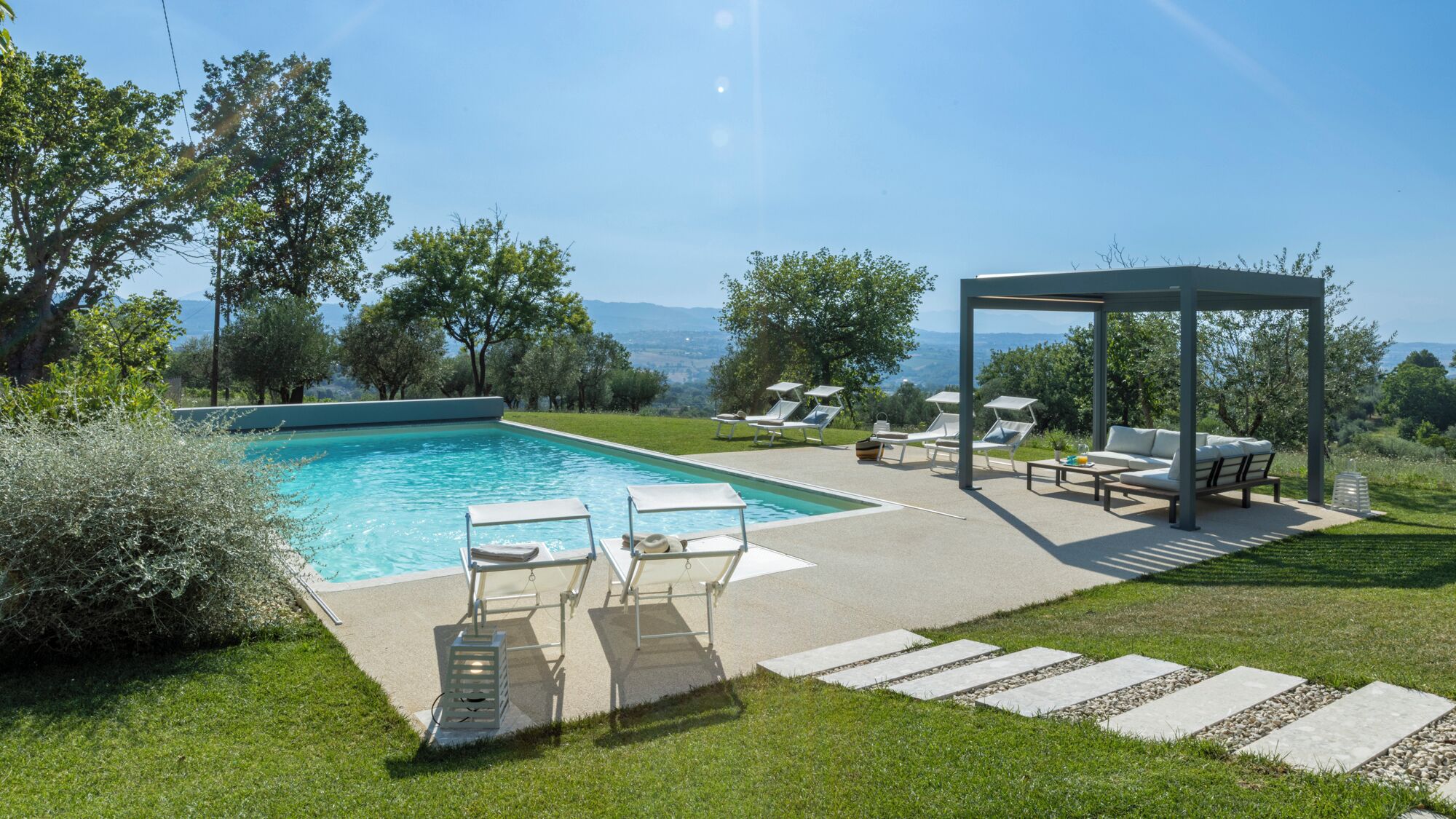 A rectangular swimming pool with a clear blue water, surrounded by a grassy area with lounge chairs and a pergola.