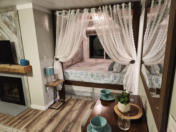 A cozy bedroom with a bed, a nightstand, a lamp, and a window with sheer curtains.