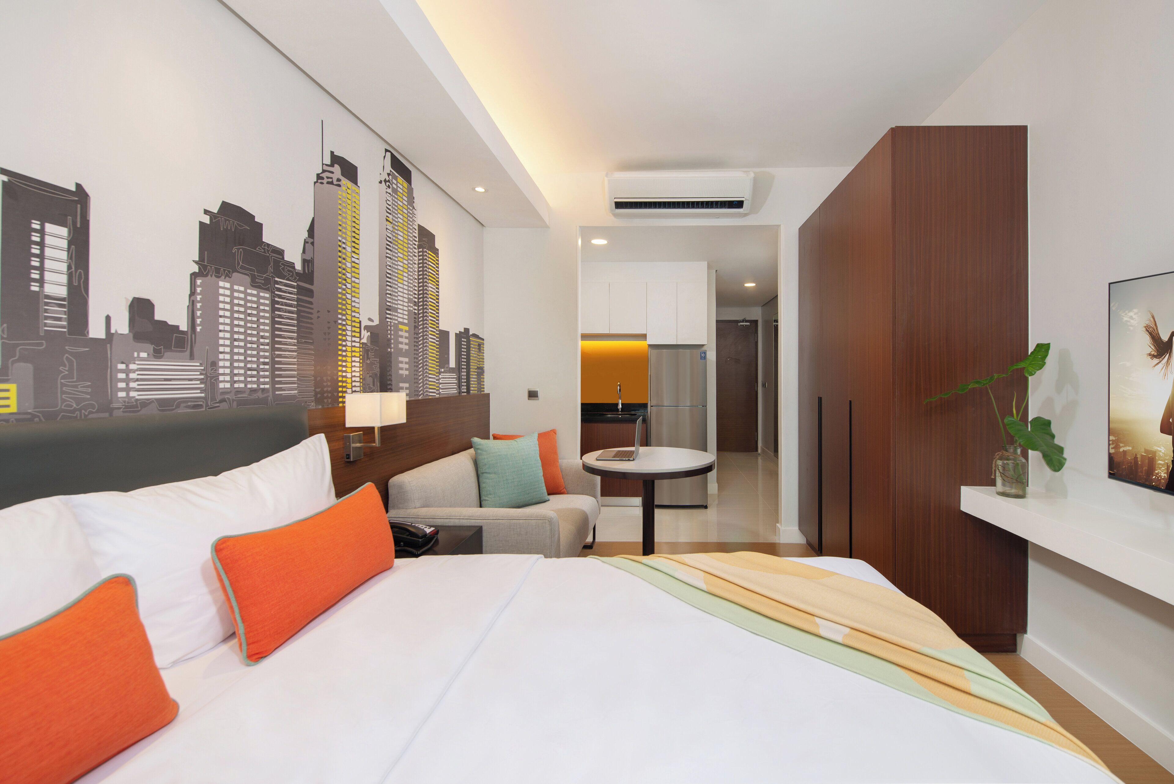 A modern hotel room with a bed, a sofa, a dining area, and a cityscape mural on the wall.