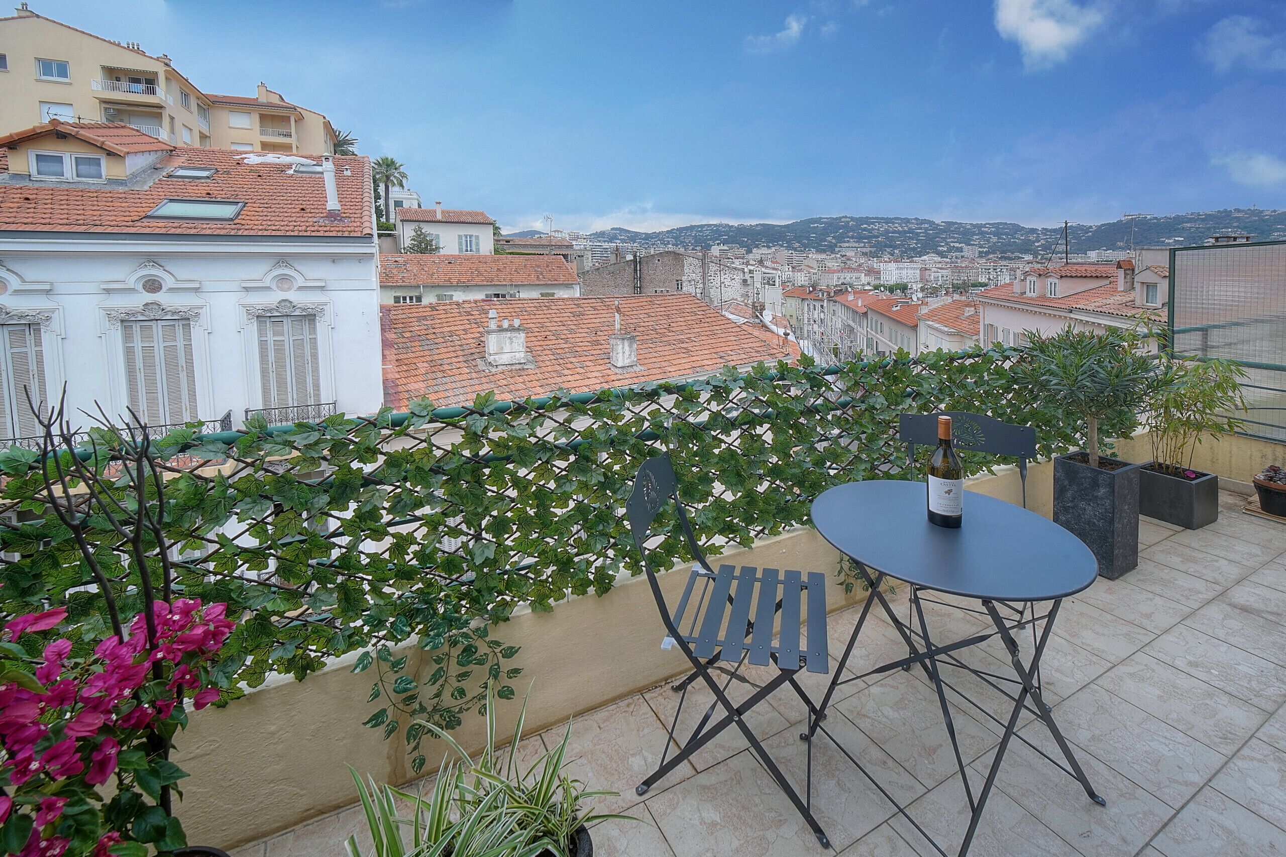 A terrace with a table and chairs, a bottle of wine, and potted plants.