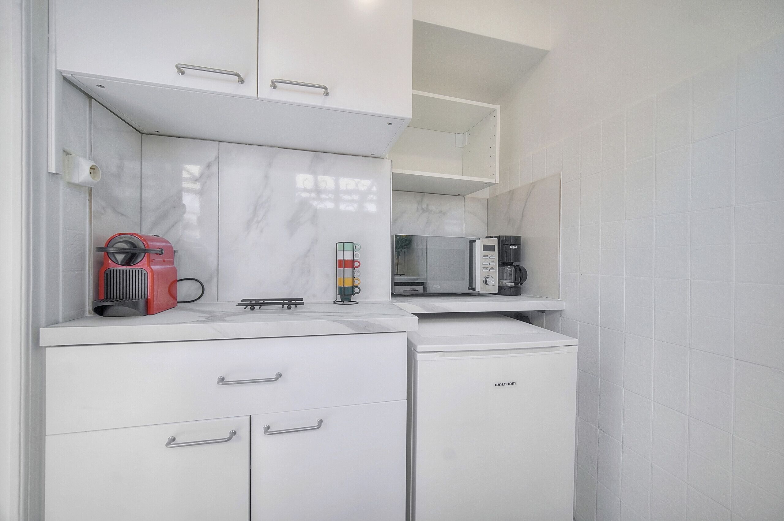 Cocina compacta con armarios blancos, microondas, cafetera y un electrodoméstico rojo en la encimera.