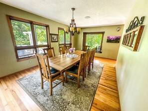 A dining room with a wooden table, chairs, and a chandelier. There are large windows with views of greenery outside.