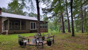 A rustic cabin with a porch, surrounded by tall trees and a fire pit with chairs.