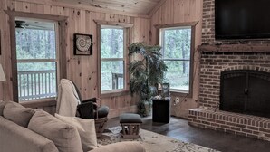 A cozy cabin interior with a wooden ceiling, a brick fireplace, a beige sofa, and large windows offering a view of the forest.