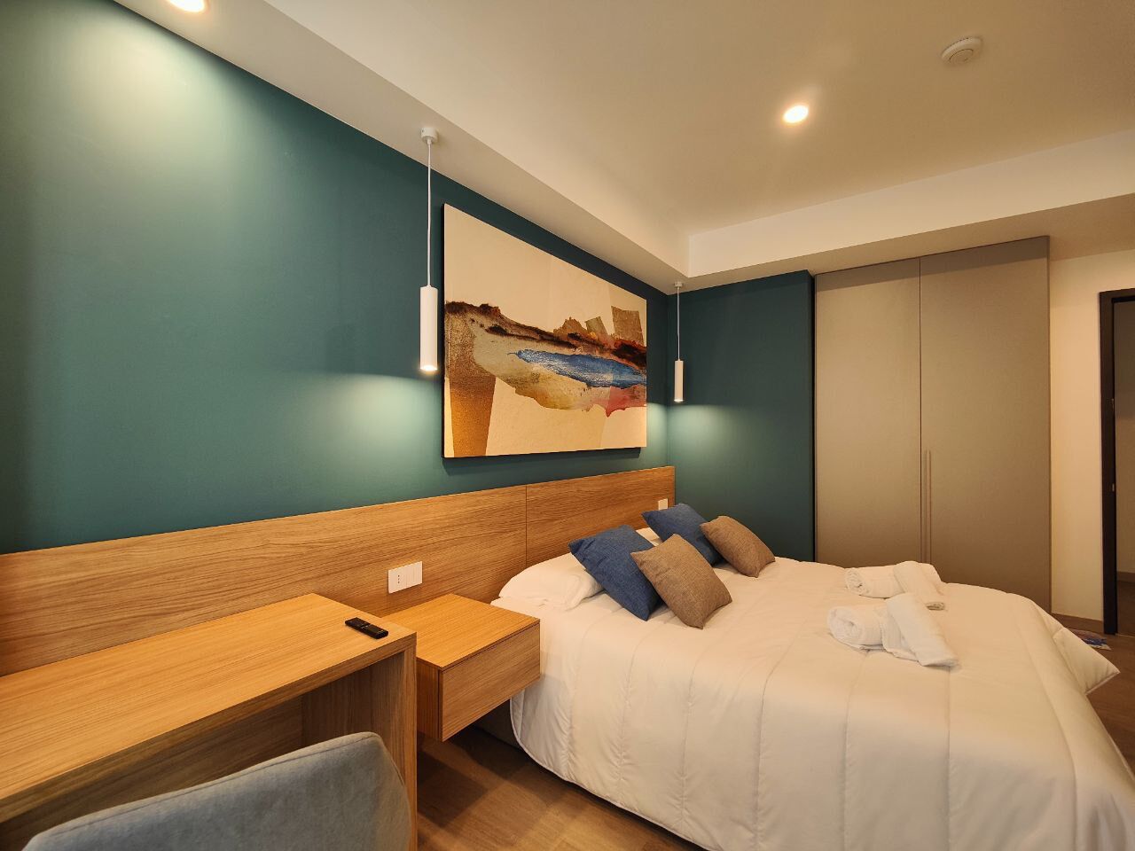 A modern hotel room with a wooden headboard, a large bed with white bedding, a desk with a chair, and a painting on the wall.