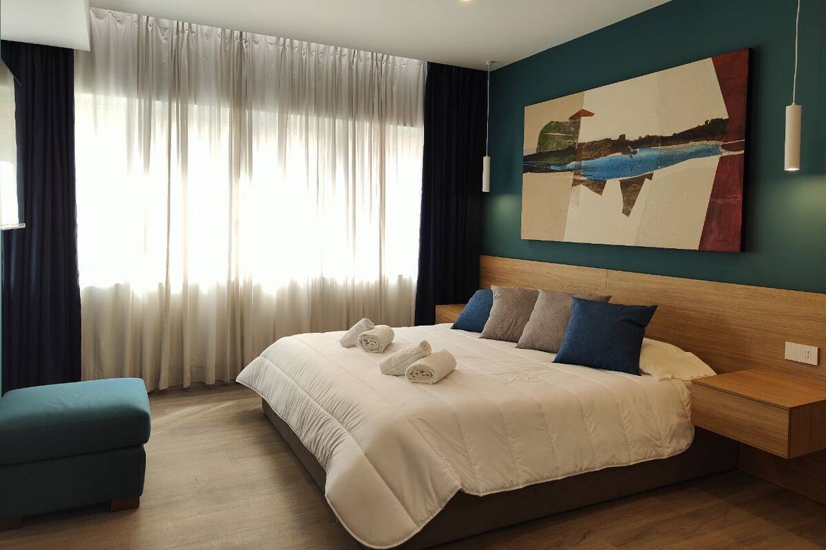 A modern hotel room with a large bed, a headboard, a bedside table, a bench, and a painting on the wall.