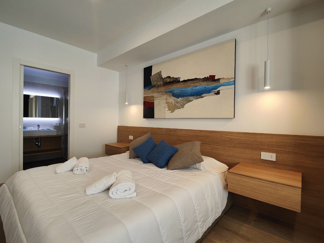 Ein modernes Hotelzimmer mit einem großen Bett, einem Holzkopfteil, einem Gemälde über dem Bett und einem Nachttisch mit einer Lampe.