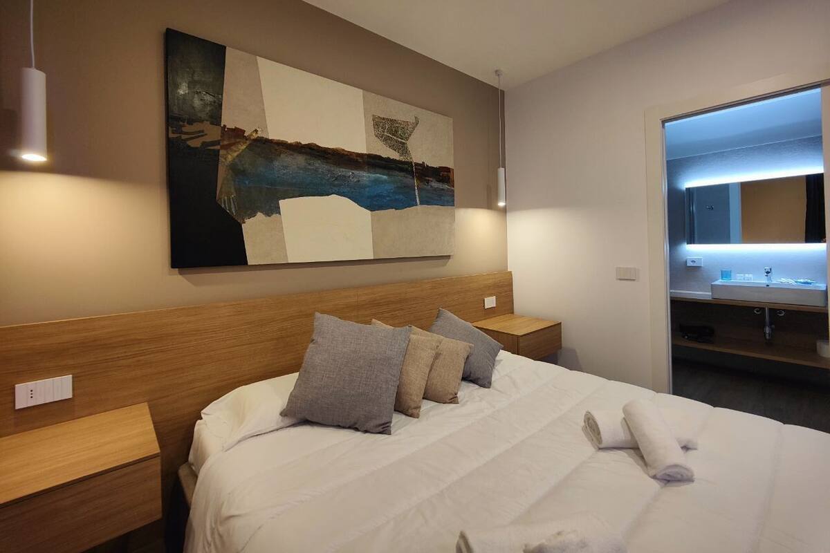 A modern hotel room with a large bed, wooden headboard, and a painting above the bed. There is a nightstand with a lamp and a bathroom visible through an open door.