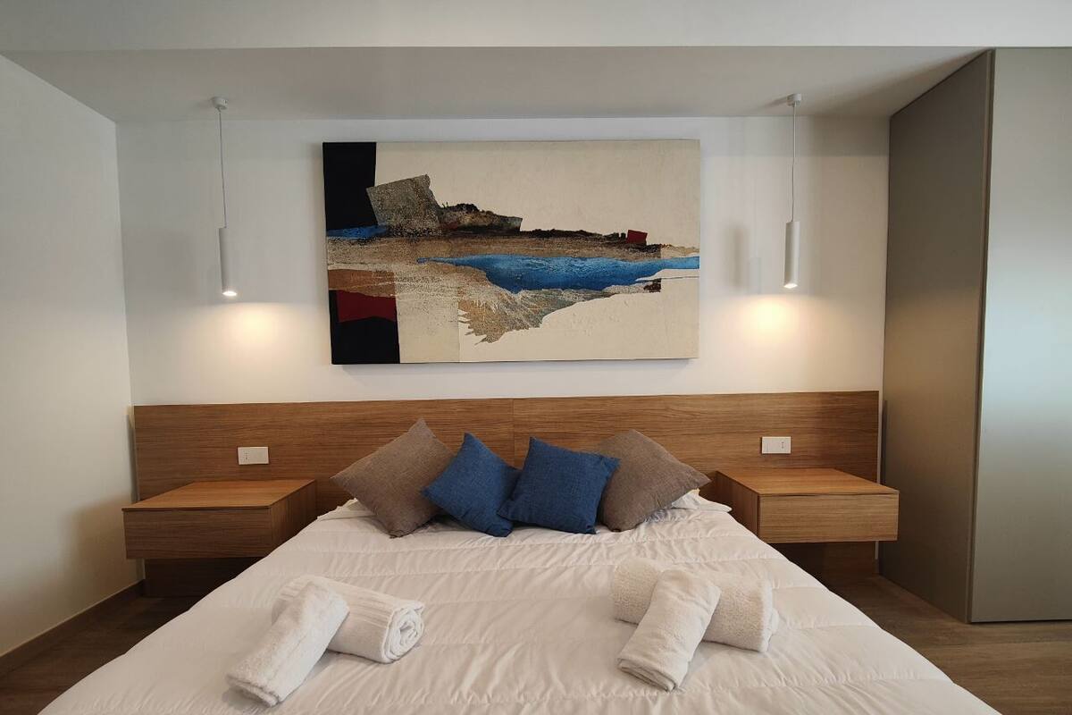 A modern hotel room with a large bed, two bedside tables, and a painting of a coastal scene.