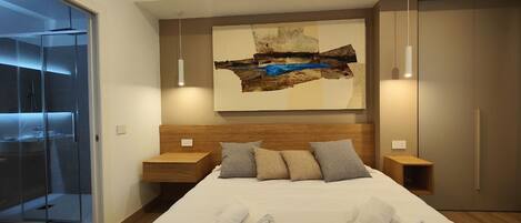 A modern hotel room with a large bed, a wooden headboard, a painting above the bed, and a sliding door leading to a shower area.
