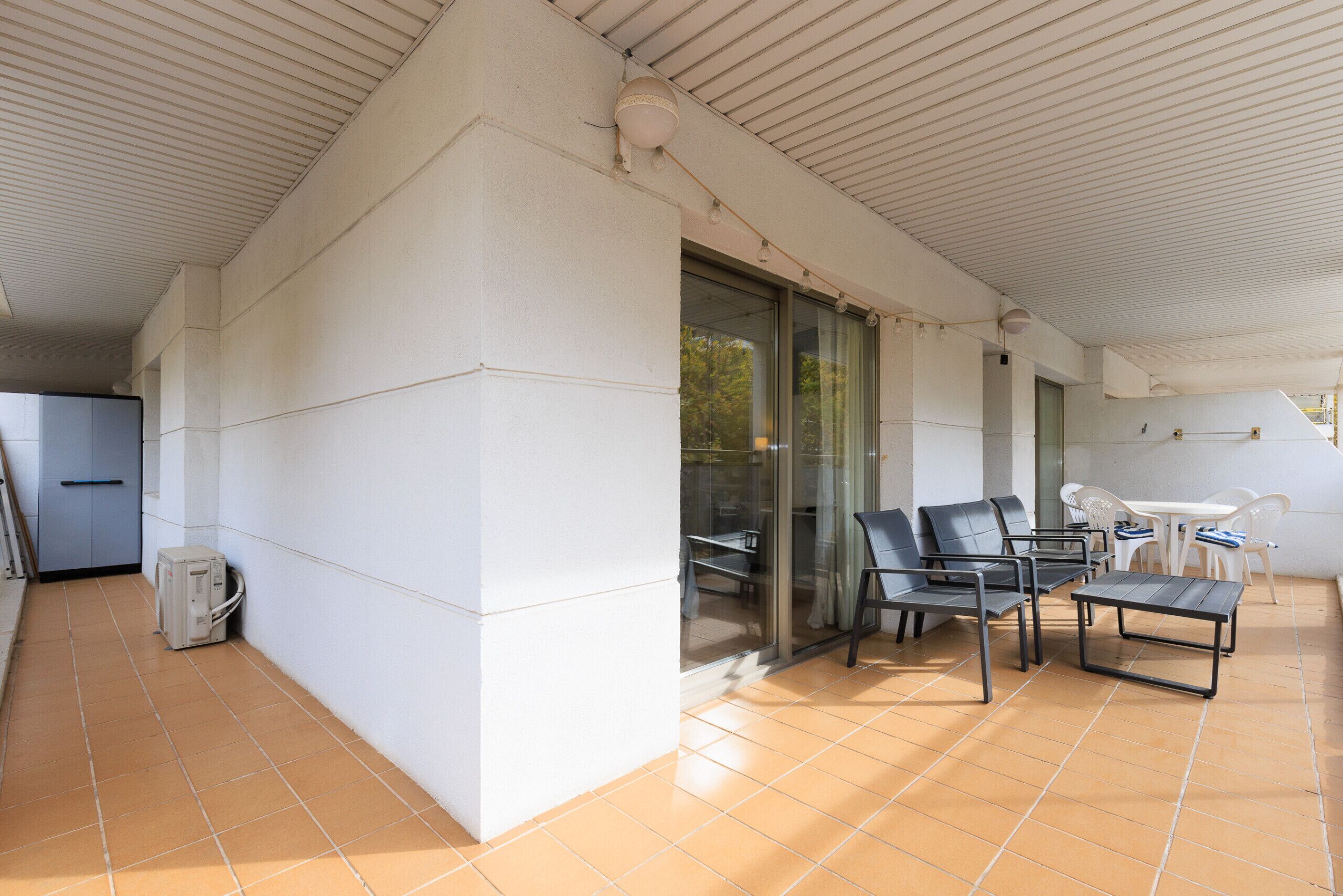 A covered outdoor area with a tiled floor, a glass door leading inside, and a seating arrangement with chairs and a table.