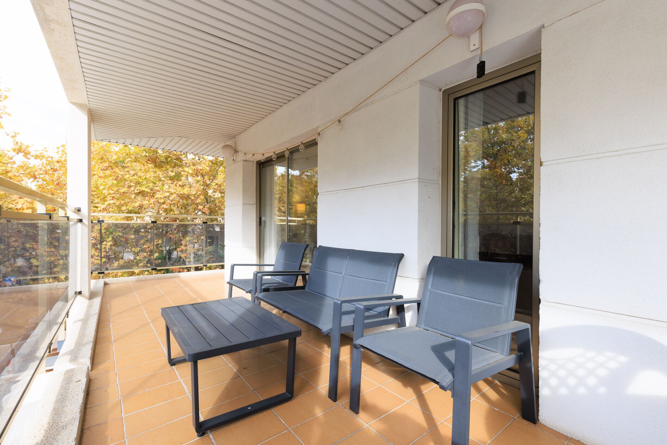 A covered patio with a table and three chairs, a glass door, and a view of trees.