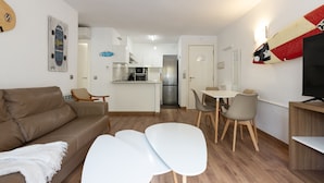 A modern living room with a brown sofa, white chairs, and a small table. A kitchen is visible in the background with a refrigerator and microwave.
