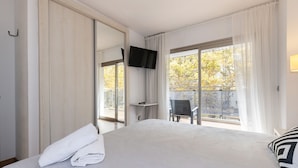 A hotel room with a large bed, a flat-screen TV mounted on the wall, a balcony with a chair, and a view of trees outside.